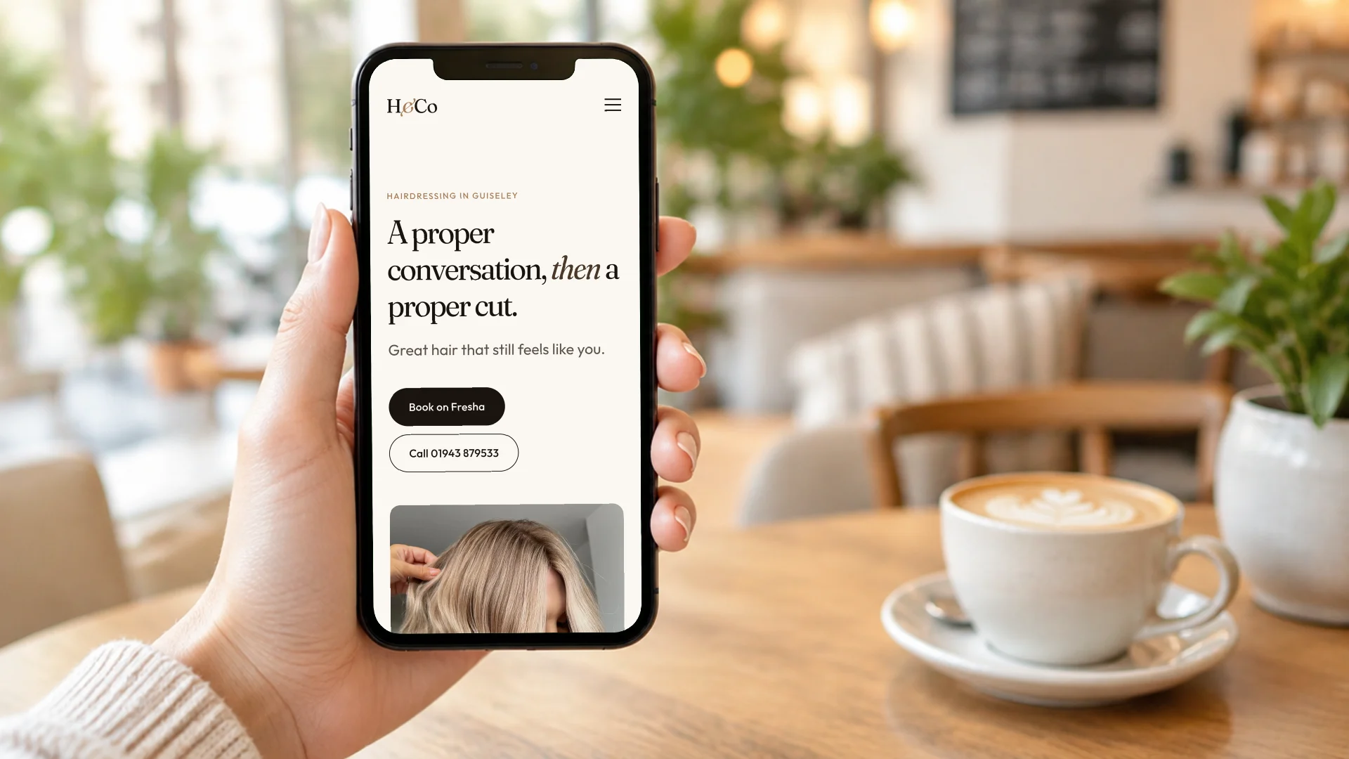 A hand holding a phone in a café showing the H&Co Hairdressing homepage, with the Book on Fresha and call buttons above the fold